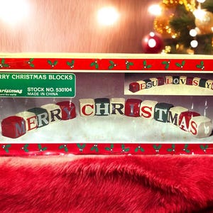 May include: A rectangular cardboard box labeled MERRY CHRISTMAS BLOCKS containing a set of multi-colored wooden blocks. The box features a red border with green mistletoe illustrations and displays the words MERRY CHRISTMAS and JESUS LOVES YOU.