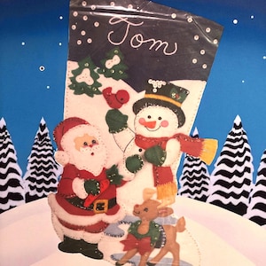 May include: A felt Christmas stocking featuring Santa Claus, a snowman, and a reindeer. The stocking is decorated with trees and a bird, with the name "Tom" written at the top. The background is a snowy landscape with a blue sky.
