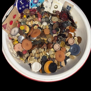 May include: A large white bowl with a red rim filled with a diverse collection of loose sewing buttons in various sizes, shapes, and colors, including brown, black, blue, and yellow, alongside several original cardboard display cards.