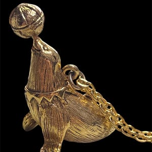 May include: A gold-toned metal pendant shaped like a seal balancing a ball on its nose. The seal has a textured surface and wears a collar with a pointed edge. A gold-colored metal chain is attached to a jump ring on the back of the seal figure.
