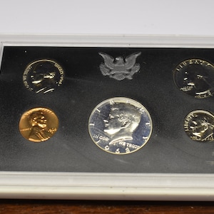 May include: A 1965 United States proof set of coins, including a half dollar, quarter, dime, nickel, and penny, displayed in a clear plastic case with a black insert. The coins are silver and copper-colored.