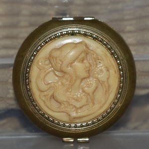 May include: A vintage compact mirror with a gold-toned metal case. The center features a cream-colored cameo of a woman's profile. The compact has a beaded border and a hinged lid, showcasing a classic, antique design.