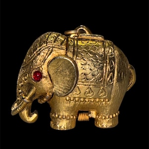 May include: A gold-toned metal elephant pendant featuring a textured finish, a small red stone eye, and intricate engraved patterns on the head and back. The elephant is depicted in profile with a trunk curved downward and a small loop at the top.