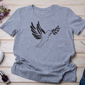 以下が含まれることがあります： オリーブの枝を持ったハトの黒いシルエットがプリントされたグレーのTシャツ。Tシャツは、白いスニーカー、マニキュアボトル、サングラスなどと一緒に木製の表面に平らに置かれています。