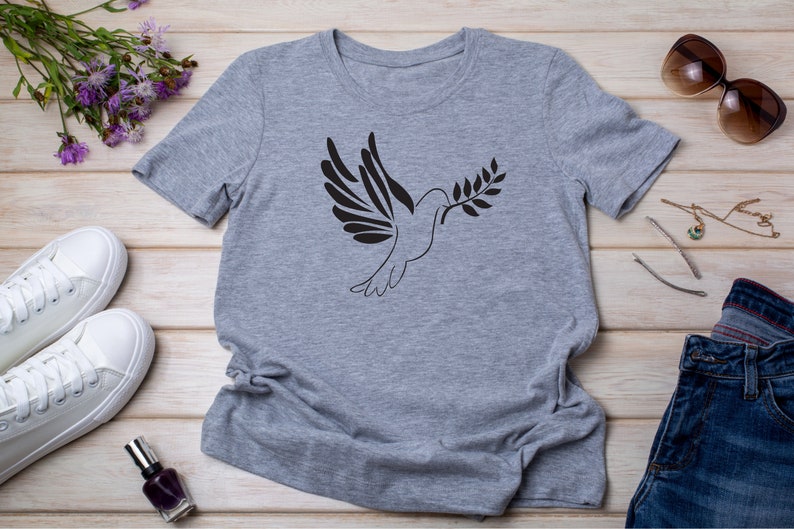 以下が含まれることがあります： オリーブの枝を持ったハトの黒いシルエットがプリントされたグレーのTシャツ。Tシャツは、白いスニーカー、マニキュアボトル、サングラスなどと一緒に木製の表面に平らに置かれています。