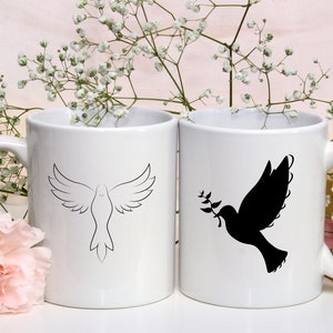 以下が含まれることがあります： 黒い輪郭の鳩が描かれた白いセラミック製のマグカップが2つ。1羽の鳩は羽を広げて飛んでおり、もう1羽の鳩はくちばしでオリーブの枝を持っています。