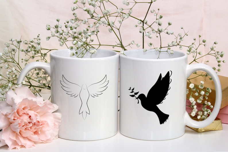 以下が含まれることがあります： 黒い輪郭の鳩が描かれた白いセラミック製のマグカップが2つ。1羽の鳩は羽を広げて飛んでおり、もう1羽の鳩はくちばしでオリーブの枝を持っています。