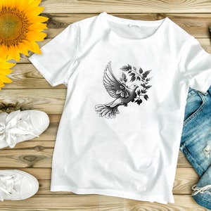 以下が含まれることがあります： 白いTシャツに、オリーブの枝を持ったハトの白黒のグラフィックがプリントされています。Tシャツは、白いスニーカーと青いジーンズと一緒に、木製の表面に平らに置かれています。