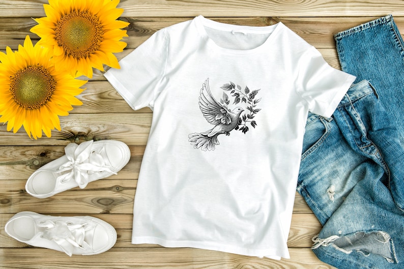 以下が含まれることがあります： 白いTシャツに、オリーブの枝を持ったハトの白黒のグラフィックがプリントされています。Tシャツは、白いスニーカーと青いジーンズと一緒に、木製の表面に平らに置かれています。