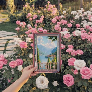 May include: A framed painting depicting a scenic landscape with a lake, mountains, and buildings, held in front of a backdrop of pink and white roses. The painting's frame is a reddish-brown color.