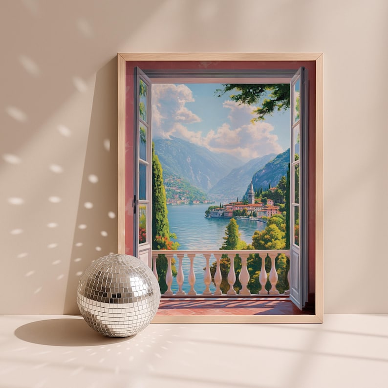 May include: A framed landscape painting depicts a scenic view through an open window. The artwork showcases a serene lake surrounded by mountains and buildings. A silver disco ball sits in front of the frame, reflecting light.