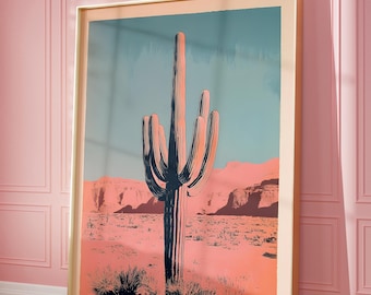 May include: Framed art print featuring a stylized illustration of a desert landscape with a large saguaro cactus. The artwork uses a palette of pinks, blues, and oranges, with the cactus in dark tones. The print is in a light-colored wooden frame.
