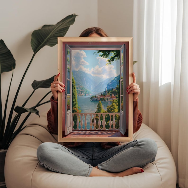 May include: A framed artwork depicting a scenic landscape viewed through an open window. The artwork features a lake surrounded by mountains, trees, and buildings. The frame is a light brown color.