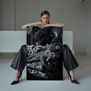 May include: Black and white artwork featuring a leopard resting on a patterned sofa. The artwork is held by a person wearing black pants and heels. The background is a neutral, minimalist space.
