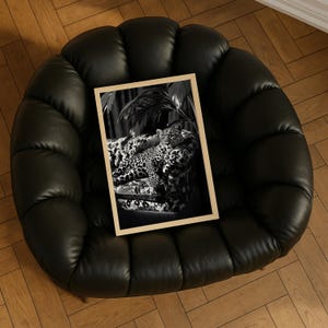 May include: A framed black and white print featuring a leopard resting on a patterned sofa. The print has a light wood frame and is set against a black leather chair. The image evokes a sense of luxury and wildlife.