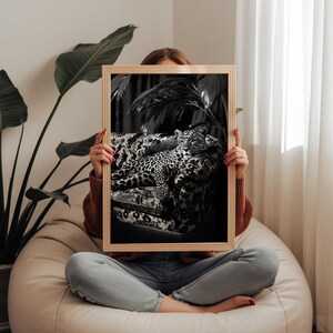 May include: A framed black and white print featuring a leopard resting on a patterned sofa. The print is held by a person sitting on a beanbag chair. The print has a light wood frame.