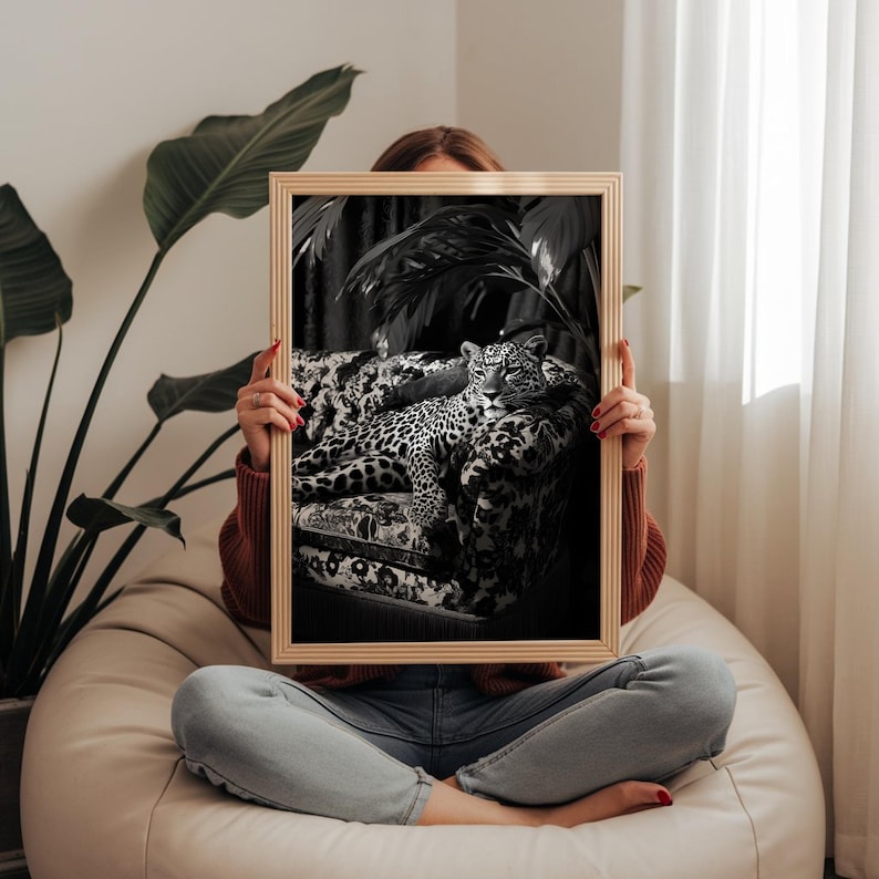 May include: A framed black and white print featuring a leopard resting on a patterned sofa. The print is held by a person sitting on a beanbag chair. The print has a light wood frame.