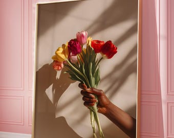 May include: A framed art print featuring a bouquet of colorful tulips. The flowers, in shades of red, pink, and yellow, are held by a hand with dark skin. The artwork is set against a neutral background and a pink wall.