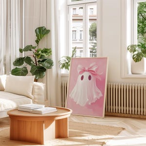 May include: A pink and white ghost with a pink bow on its head. The ghost is on a pink background. The image is in a wooden frame.