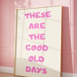 Pode incluir: Uma impressão artística emoldurada com as palavras "THESE ARE THE GOOD OLD DAYS" em letras rosa e em negrito. A obra de arte é definida contra um fundo creme e emoldurada em uma moldura dourada. A impressão está encostada em uma parede rosa.