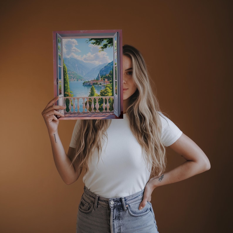May include: A framed painting depicting a scenic view through an open window. The artwork showcases a serene landscape with a lake, mountains, and buildings. The frame is a light brown color, and the painting has a detailed, realistic style.