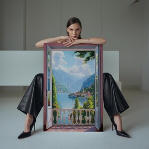 May include: A framed painting depicting a scenic landscape with a lake, mountains, and a village. The painting is held by a person wearing black pants and heels. The frame is a muted red color.