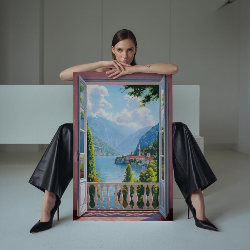 May include: A framed painting depicting a scenic landscape with a lake, mountains, and a village. The painting is held by a person wearing black pants and heels. The frame is a muted red color.