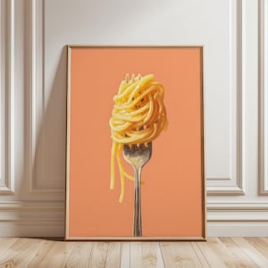 May include: A framed print featuring a close-up of spaghetti twirled on a silver fork. The pasta is a golden yellow color, and the background is a solid peach color. The print is displayed on a wooden floor.
