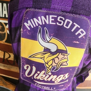 May include: Purple and black plaid flannel shirt with a patch featuring the Minnesota Vikings logo and the word "Vikings". The patch has a yellow stripe and the words "Minnesota" and "Football".