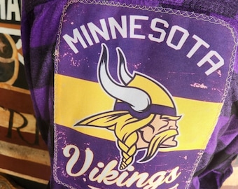 May include: Purple and black plaid flannel shirt with a patch featuring the Minnesota Vikings logo and the word "Vikings". The patch has a yellow stripe and the words "Minnesota" and "Football".