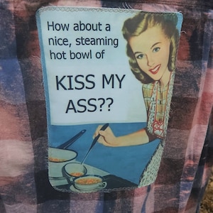 May include: A plaid shirt with a patch featuring a vintage illustration of a woman and the text "How about a nice, steaming hot bowl of KISS MY ASS??" The image shows a woman smiling and cooking soup.