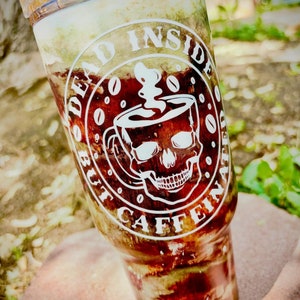 Op de afbeelding: Een witte tumbler met een schedel en gekruiste beenderen ontwerp en de tekst "Dead Inside But Caffeinated". De tumbler is gevuld met een bruine vloeistof en heeft een wit deksel.