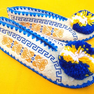 以下が含まれることがあります： 青と金の刺繍が施された白いスリッパのペア。スリッパは青いトリムがあり、幾何学模様が繰り返されています。各スリッパのかかとには、青、黄、白のポンポンが付いています。