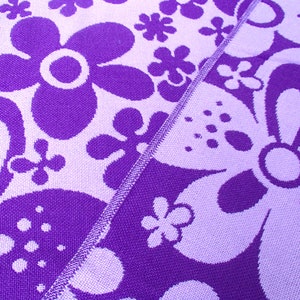 May include: Purple and white fabric with a floral pattern. The fabric has a textured surface and a repeating pattern of flowers.