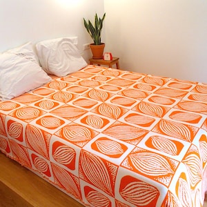 Puede incluir: Una cama blanca con una colcha de estampado naranja y blanco. La colcha tiene un diseño geométrico con líneas blancas sobre un fondo naranja.
