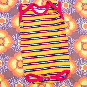 Puede incluir: Un body de bebé a rayas amarillas y rojas. El body tiene rayas horizontales en amarillo, azul y rojo. El ribete alrededor del cuello y las piernas es rojo. El fondo es un estampado floral marrón y naranja.