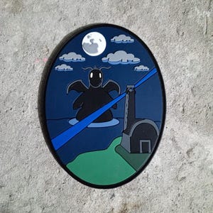 May include: A blue and green circular patch with a black silhouette of a creature with wings and a white moon in the background. The creature is standing in the water with a blue beam of light shining from a tower on the right side of the patch.