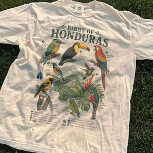 May include: Ivory-colored t-shirt featuring a colorful illustration of various birds native to Honduras. The design includes the text "Birds of Honduras" in green, along with the word "Ivory" at the bottom. The shirt is laid on a bed of green grass.