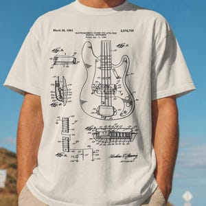 Puede incluir: Camiseta de color crema con un diagrama en blanco y negro de una guitarra el&eacute;ctrica. La camiseta incluye texto que indica una fecha de patente del 28 de marzo de 1961. La persona de la imagen lleva una gorra de b&eacute;isbol beige.