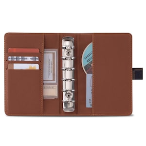 May include: A brown leather bound planner with a six-ring binder system. The planner has a card slot, a pocket for notes, and a clear pocket for a business card or other small item. The planner is open to reveal the inside, which has a silver binder mechanism.