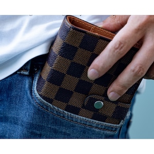 May include: A brown and black checkered wallet with a silver button. The wallet is in a blue jean pocket.