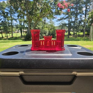May include: A transparent red castle model with two towers and a central structure, placed on a black cooler with cup holders. The scene is set outdoors with a green lawn and trees in the background.