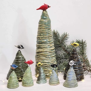 May include: A collection of ceramic tree sculptures in various sizes and colors. Each tree is topped with a small, colorful bird figurine. The trees have a textured, layered design with shades of green, blue, and brown. A red cardinal sits atop the tallest tree.