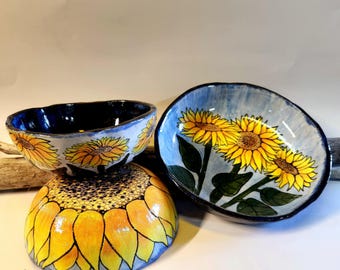 May include: Three ceramic bowls with sunflower designs. The bowls feature yellow sunflowers with black and green details against a blue background. The bowls are hand-painted and have a unique, organic shape. The bowls are suitable for serving food or as decorative items.