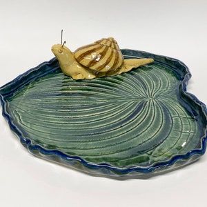 May include: A ceramic dish shaped like a leaf with a blue and green glaze. A small ceramic snail is sitting on the leaf.
