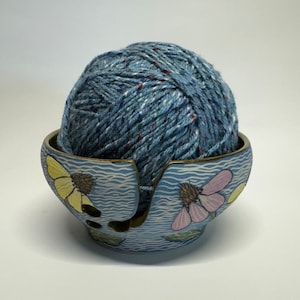May include: A blue ceramic yarn bowl with a floral design. The bowl has two openings for yarn to feed through. A ball of blue yarn is sitting in the bowl.