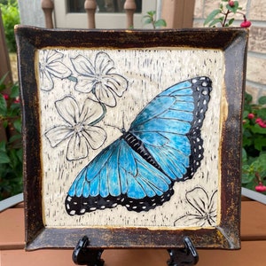May include: A square ceramic plate with a dark brown border and a white background. The plate features a blue butterfly with black markings and white flowers.