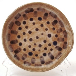 May include: A handmade ceramic plate with a speckled tan background. The plate features a pattern of dark brown rectangles and dots. The rim is a darker shade of brown. The plate is on a clear plastic stand.