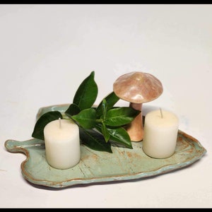 May include: A ceramic leaf-shaped tray in a muted teal color, holding two white candles, green leaves, and a ceramic mushroom. The tray has a textured surface and a brown rim, creating a rustic aesthetic.