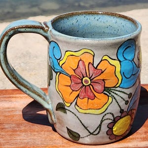 May include: A handmade ceramic mug with a light gray exterior and a blue interior. The mug is decorated with vibrant orange, red, and blue floral designs. The handle and rim have a brown glaze. A unique piece for enjoying beverages.